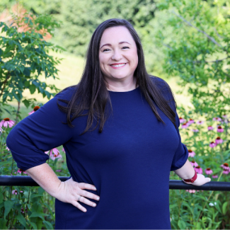Portrait of Julianna Jones, a woman with long dark hair, smiling in a navy blue dress in front of a garden with purple flowers.
