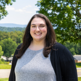 Julie Tilson smiling outdoors, wearing a gray heathered top under a black cardigan, with trees and a distant mountain landscape in the background.