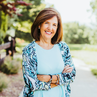 Professional headshot of Karen Cheers, a smiling woman with brown hair, wearing a light blue top and a white cardigan with a blue floral pattern, standing outdoors on a paved path.