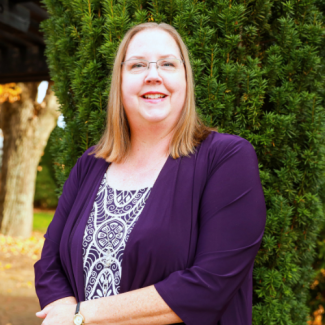 Headshot of Karen Viers, a woman with blonde hair and glasses, smiling in a purple cardigan over a patterned top. She is standing outdoors in front of a tall evergreen.