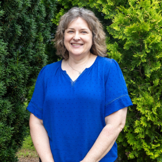 Headshot of Karin Widener, a woman with wavy light brown hair, smiling in a bright blue blouse. She is standing outdoors between two green evergreen trees.