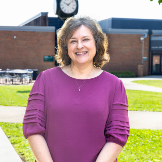 Karin Widener standing outside on a campus courtyard, wearing a purple blouse and smiling.