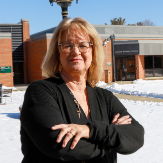 Headshot of Kathy Smith, a woman with blonde hair and glasses, posing with arms crossed. She is standing outdoors in a snowy courtyard in front of the Collins Student Center.