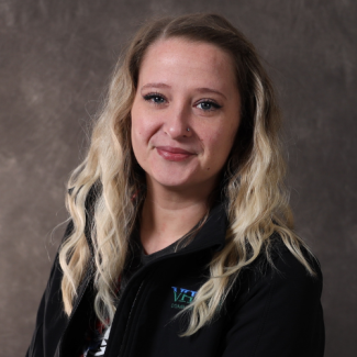 Professional headshot of Katie Applegate, a woman with blonde, wavy hair, wearing a dark jacket, against a neutral gray background.