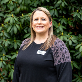 A woman with long blonde hair and a friendly smile poses in front of a dense green rhododendron bush. She wears a black top with grey animal-print panels on the shoulders and a "Virginia Highlands" nametag that identifies her as Kayla Mullins.