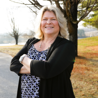 Professional headshot of Kelly Emery, a smiling woman with blonde hair, wearing a patterned black and white top and a black cardigan, standing outside with a tree in the background.
