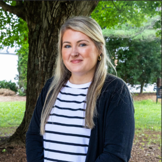 Headshot of Kenna Widener smiling outdoors in front of a tree, wearing a black-and-white horizontal striped shirt and a black cardigan.