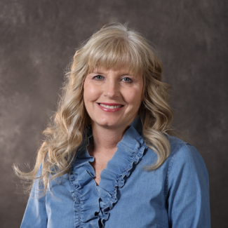 Studio portrait of Kim Felty, a woman with long blonde wavy hair and bangs, wearing a blue ruffled denim shirt.