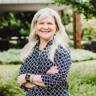 Portrait of Lee Hunt, a woman with long blonde hair, smiling with arms crossed in a garden setting.