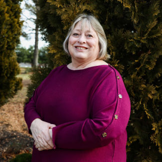 Professional headshot of Linda Colley, a smiling woman with blonde hair, wearing a magenta sweater, standing in front of evergreen trees.