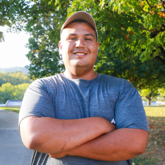 Luke Green, a man wearing a grey t-shirt and a tan baseball cap, smiling with arms crossed outdoors with a large tree in the background.