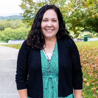 Professional headshot of Mendy Bechtold, a smiling woman with long, dark curly hair, wearing a green patterned dress and a black cardigan, standing outdoors.