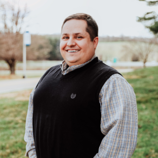 Michael McBride, a man wearing a plaid shirt and a black sweater vest, smiling in an outdoor setting with a grassy field in the background.