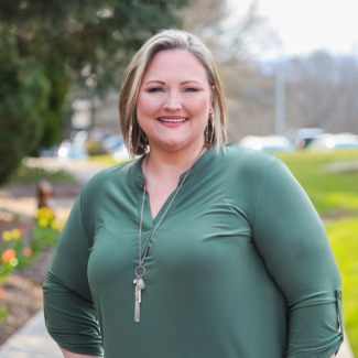 Professional headshot of Michelle Phillips, a woman with blonde hair, smiling and wearing a green top and a long necklace against an outdoor blurred background.
