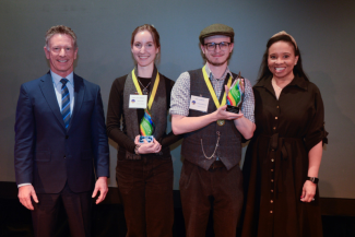 Four people stand smiling on a stage for an awards presentation. Two students in the center wear yellow medallions and hold colorful glass trophies. They are flanked by a man in a navy suit on the left and a woman in a black dress on the right.