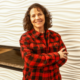 Paige Kelly, a woman with curly brown hair, wearing a red and black plaid jacket, smiling with arms crossed in front of a white wavy-textured wall.