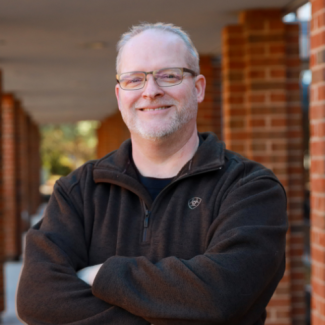 Professional headshot of Preston Ball, a smiling man with glasses and a short beard, wearing a dark brown fleece jacket, standing in a brick-lined walkway.
