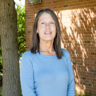 Reva Russell, a woman with long brown hair wearing a light blue sweater, smiling in front of a brick building and a tree.