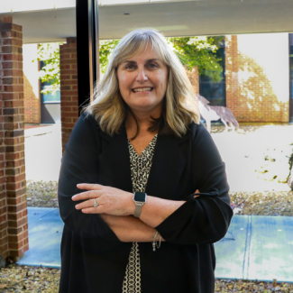 Shannon Mutter, a woman with blonde hair, wearing a patterned blouse and a black cardigan, smiling with arms crossed in front of a window showing a brick building.