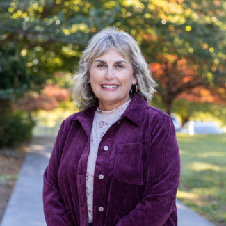 Shannon Mutter smiling, wearing a purple corduroy jacket over a patterned top outdoors with a paved walkway in the background.