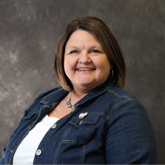 Sharon Rowe, a woman with short brown hair, wearing a white shirt and a denim jacket with a decorative pin, smiling against a dark grey textured background.