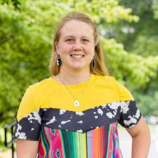 Headshot of Shasta Wilson smiling outdoors, wearing a vibrant yellow, cow-print, and multi-colored striped top.