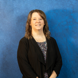 A professional headshot of Andrea Repass, a woman with shoulder-length wavy brown hair, smiling and wearing a black cardigan over a floral print blouse against a textured blue background.