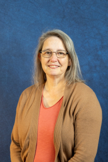 Headshot of Cindy Woosley, a smiling woman with long gray hair and glasses, wearing a coral shirt and a tan cardigan against a textured blue background.