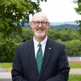 Adam Hutchison smiling in a dark grey suit jacket and green striped tie, with a soft-focus background of green trees and mountains.