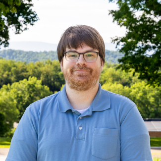 A medium close-up headshot of Joe Hamil, a man with brown hair, bangs, glasses, and a light brown beard, smiling gently. He is wearing a light blue polo shirt, set against a blurred background of green summer trees under a bright sky.
