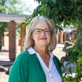 Kathy Smith smiling in a green cardigan and white blouse, framed by green leaves with a soft-focus background of an outdoor courtyard and brick pillars.
