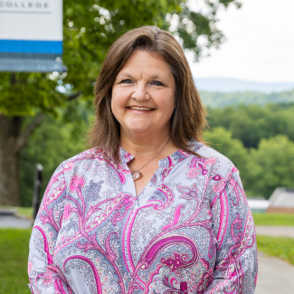 A medium close-up headshot of Sharon Rowe, a woman with shoulder-length brown hair, smiling warmly. She is wearing a pink and grey paisley patterned split-neck blouse and a necklace. The background is a blurred outdoor scene with green trees, hills, and a partial sign on the left.