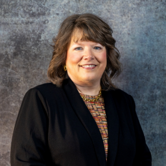 A woman with shoulder-length wavy brown hair and bangs smiles warmly. She is wearing a black blazer over a multi-colored patterned blouse, accented by a gold chain necklace and gold hoop earrings. She stands against a grey, textured studio wall.