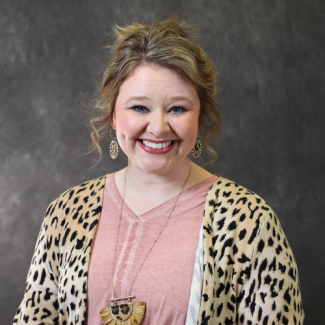 Professional headshot of Taylor Evans (Skeens), a smiling woman with blonde hair in an updo, wearing a pink top and a leopard-print cardigan, against a gray background.