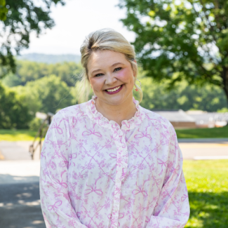 Taylor Evans smiling outdoors, wearing a white blouse with a pink floral and bow pattern and a ruffled collar, with green trees in the background.