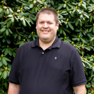 A man with short brown hair and a friendly smile stands in front of a dense green bush. He is wearing a black short-sleeved polo shirt.