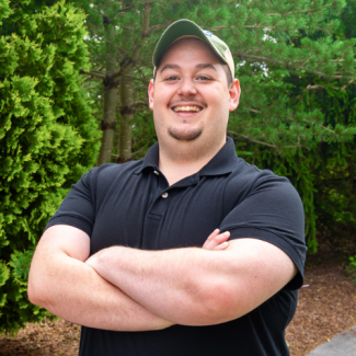Trevor Harrison, a man with a goatee wearing a black polo shirt and a green and tan baseball cap, smiling with arms crossed outdoors with green trees in the background.