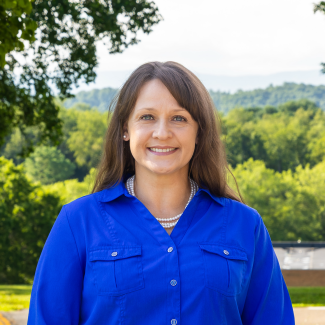Hannah Vaughn smiling in a bright blue collared shirt and pearl necklace, with a soft-focus background of green trees and hills.