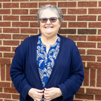 Vota Thomas, a woman with short grey hair and glasses, wearing a blue patterned shirt and dark blue cardigan, smiling in front of a brick wall.