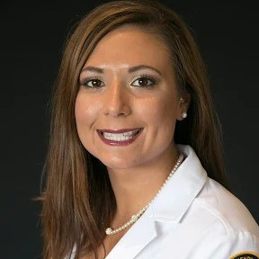 Professional headshot of Whitney Peters, wearing a white lab coat and a pearl necklace, smiling against a black background.