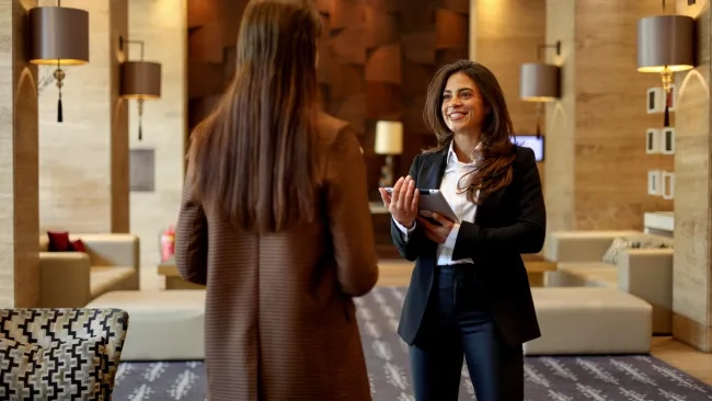 Two professional women converse in a modern, well-lit hotel lobby. One woman with long brown hair stands with her back to the camera, wearing a brown patterned trench coat. Facing her is a smiling woman with dark hair, dressed in a black blazer, white button-down shirt, and dark pants, holding a silver tablet. The background features upscale hotel decor with soft lighting, wall lamps, plush seating, and textured walls.