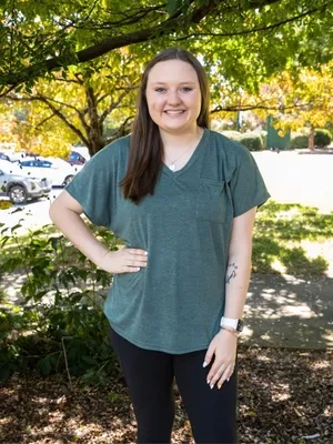 A smiling young woman with long brown hair poses outdoors, wearing a green v-neck t-shirt and black pants. She is standing in front of lush green and yellow autumn trees.