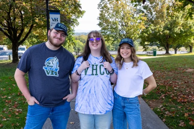 Three smiling students standing together on a campus path. The male student wears a dark gray VHCC wolf logo t-shirt, the middle student wears a blue tie-dye VHCC shirt, and the female student on the right wears a white t-shirt and a green VHCC cap.